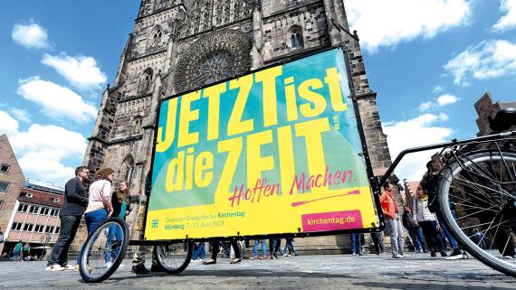 Gelbe Schrift auf grünem Grund: das Kampagnenmotiv zum Deutschen Evangelischen Kirchentag 2023 mit der Losung „Jetzt ist die Zeit“ vor der Nürnberger Lorenzkirche.